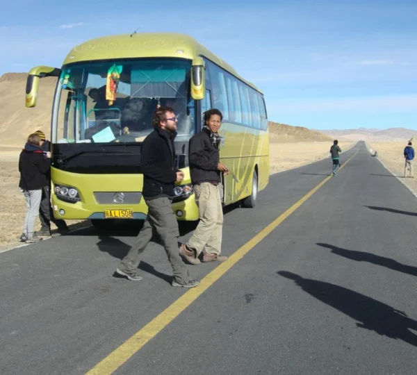 Tourists standing by a yellow tour bus on an open highway during a road trip to Tibet under a blue sky.