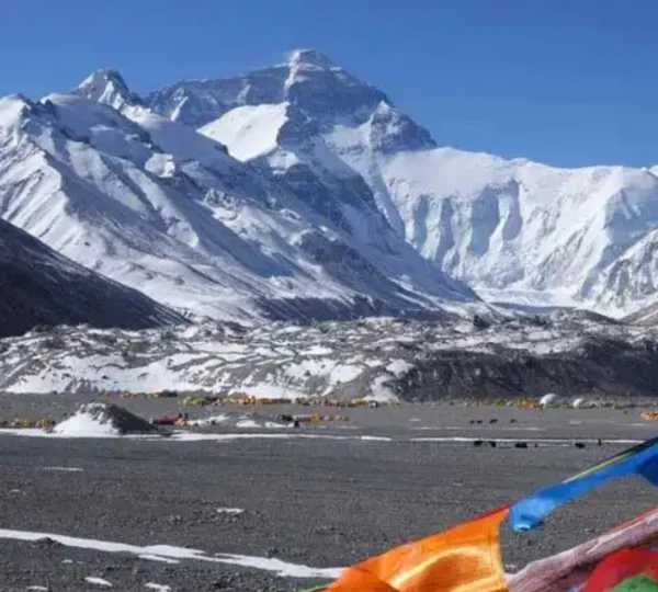 Wide view of a massive, snow-covered mountain peak rising above a gravelly valley base camp filled with distant tents and colorful prayer flags in the foreground.