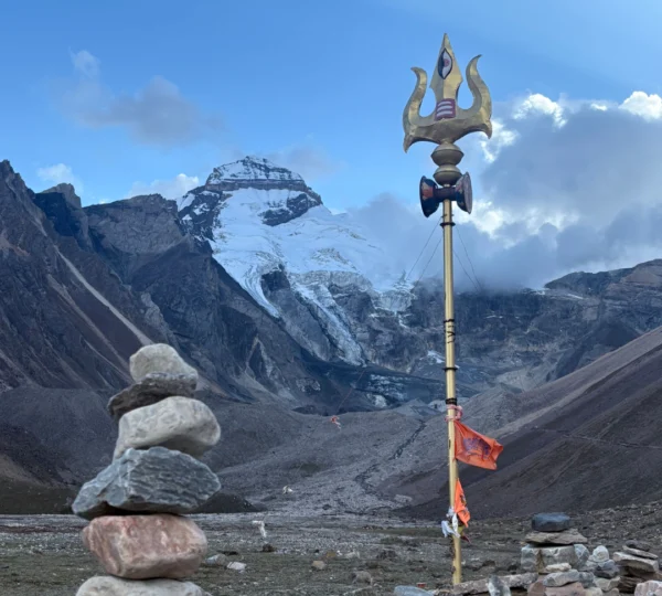 A golden Shiva Trishul (trident) adorned with a small drum and orange flags stands in a rocky valley with stacked stone cairns and a snow-dusted mountain in the background.