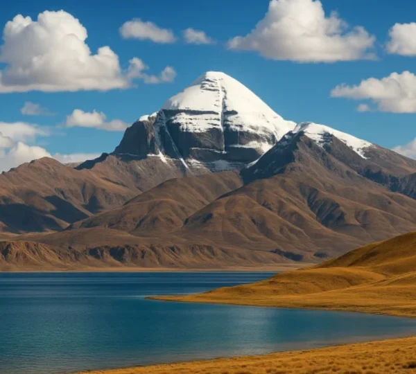 Snow-capped peak of Mount Kailash rising behind the deep blue waters of Lake Mansarovar under a blue sky with scattered white clouds.