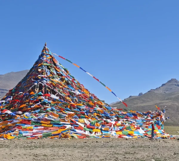 A massive, pyramid-shaped mound completely covered in thousands of colorful Tibetan prayer flags under a clear blue sky, with an arid mountain landscape behind it.